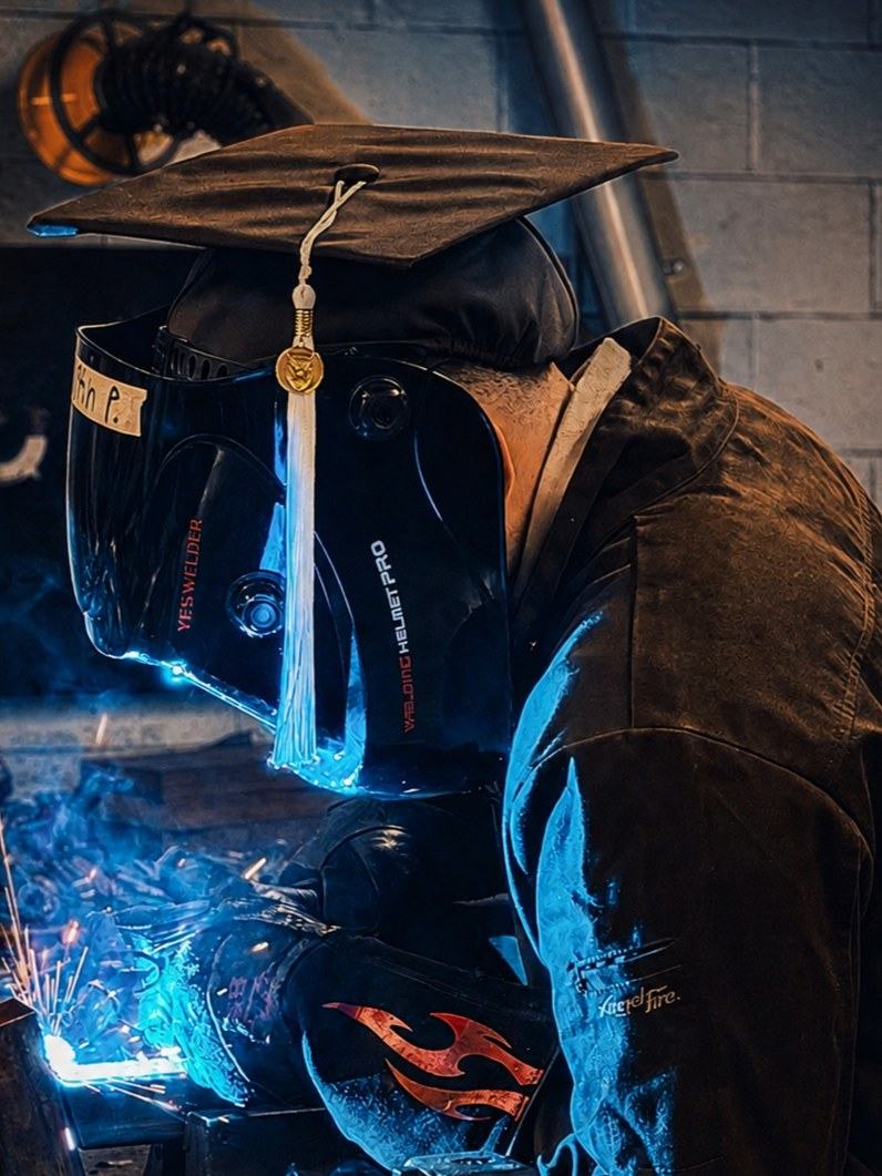 Welding in the shop wearing a graduation cap over a welding helmet